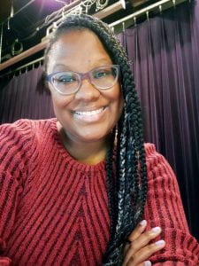 Photo of a Black woman with braided hair who is wear glasses and a red top. She is smiling in to the camera with a stage in the background.